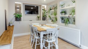A dining area with a wooden table and white chairs, a TV mounted on the wall, and shelves with potted plants. - 2 bedroom accommodation in Torquay (Torquay)