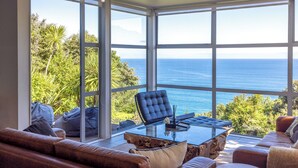 A spacious living area with a large glass window overlooking the ocean, a leather sofa, a glass coffee table, and a blue chair. - Bellevue, Onetangi - Sea Views, Modern Comfort, and Relaxation (Waiheke Island)
