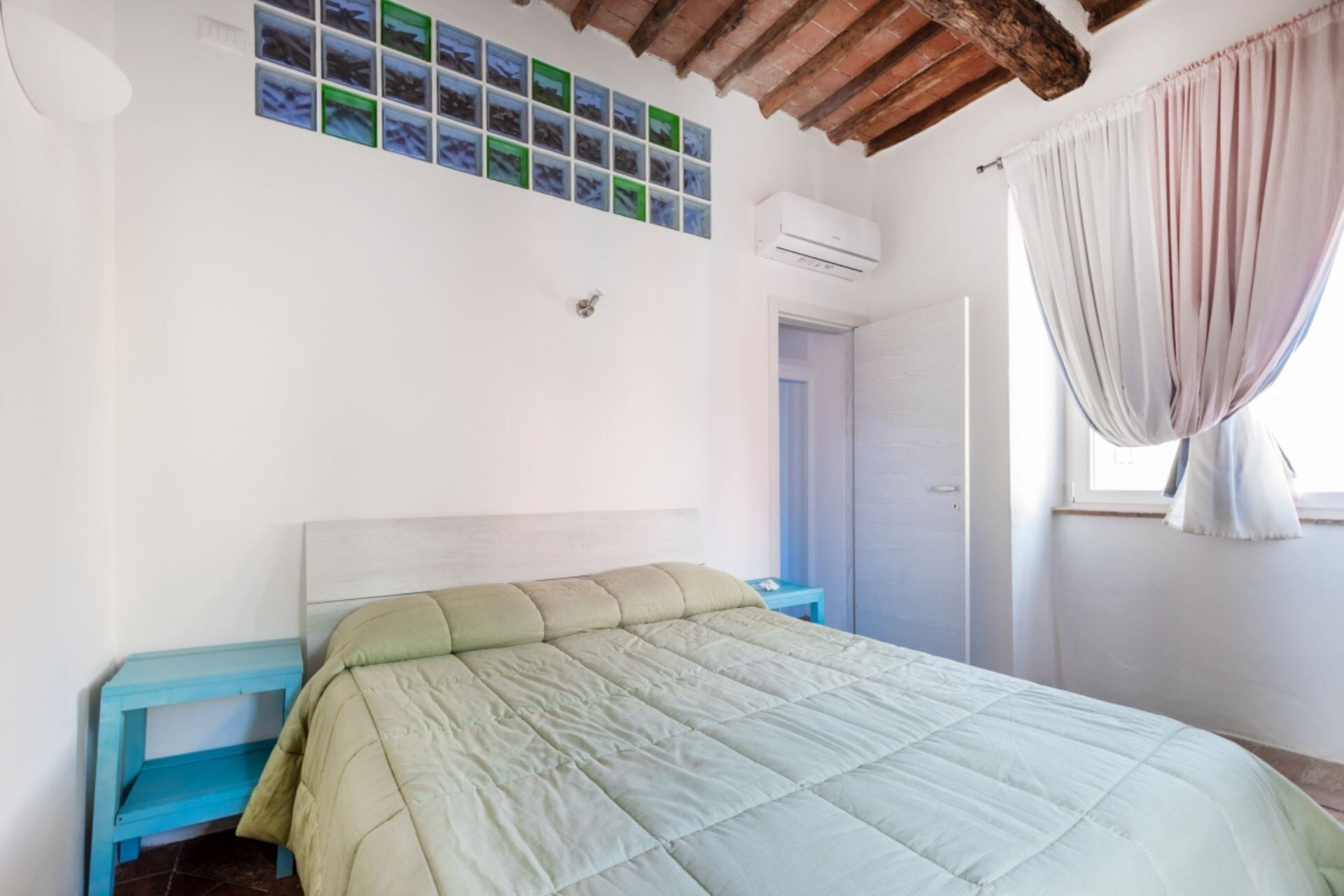 Une chambre à coucher avec un lit, une table de chevet bleue, une tête de lit blanche, un climatiseur fixé au mur, une porte et une fenêtre avec des rideaux.