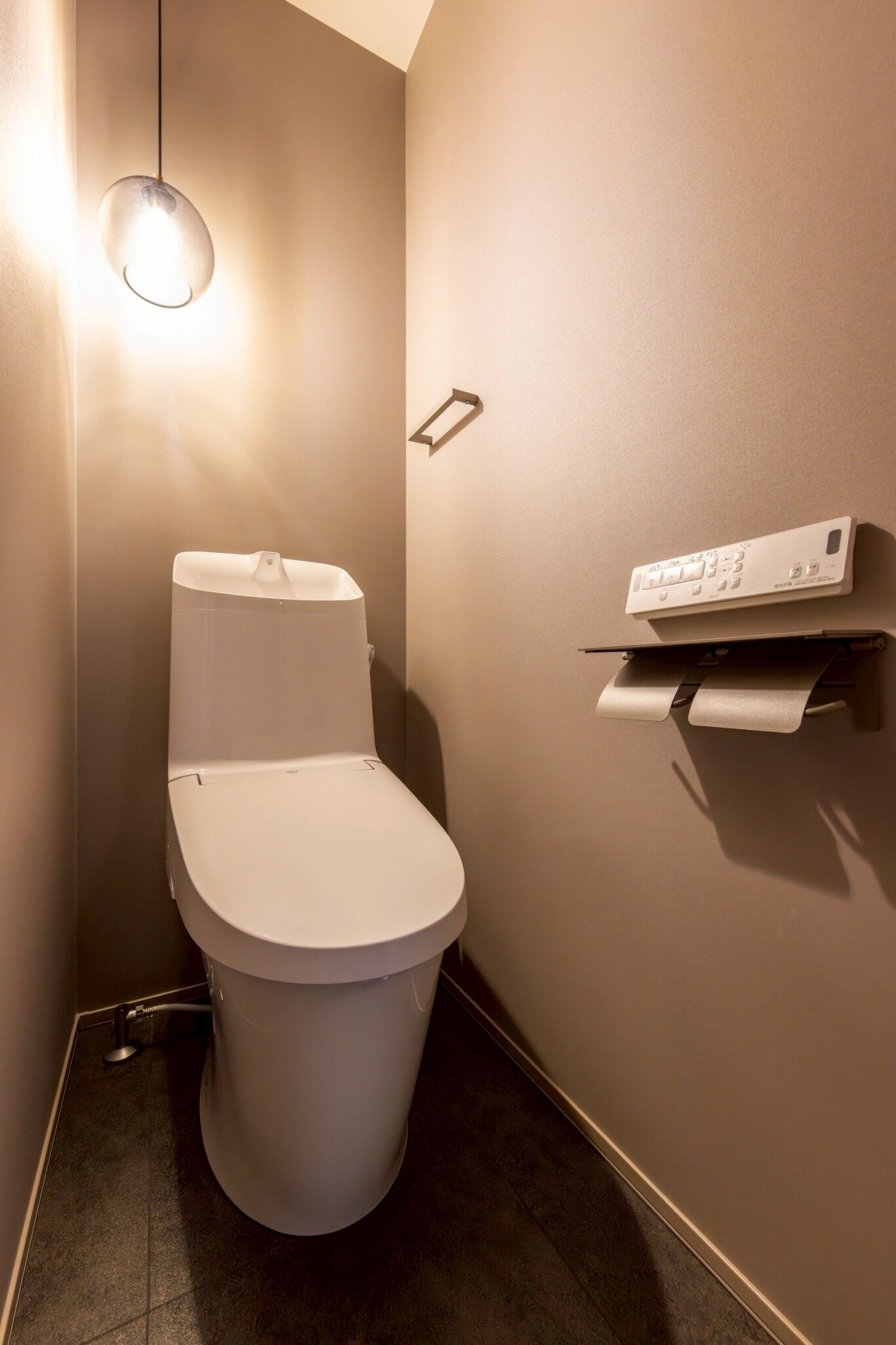 Une salle de bain moderne avec des toilettes, une étagère fixée au mur où est posé du papier toilette, et un luminaire suspendu.