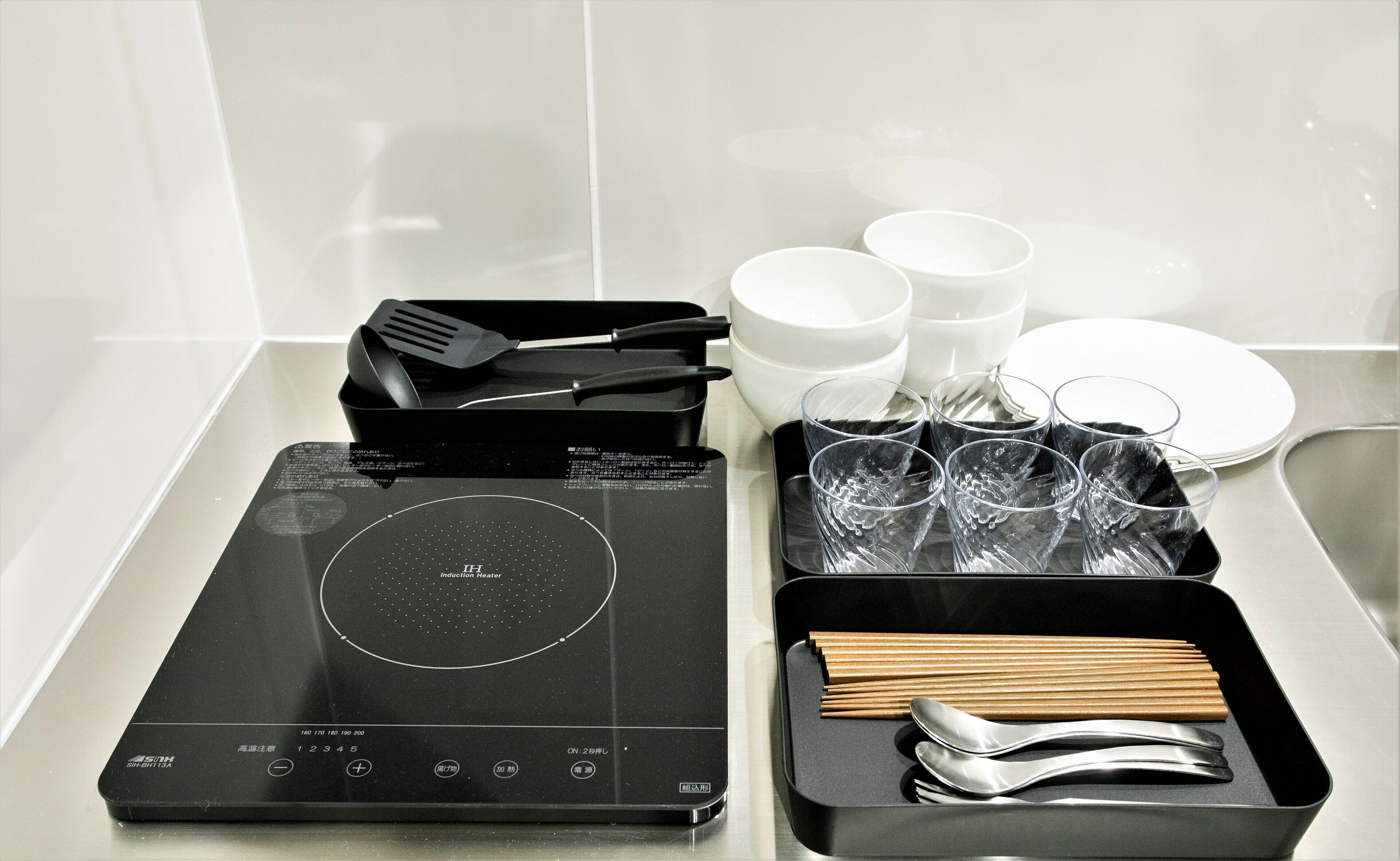 A kitchen with an induction cooktop, a black spatula, a set of wooden chopsticks, a black tray with a spoon and fork, and several white bowls and glasses.