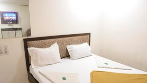 Una cama bien hecha con ropa blanca y un cabecero marrón, un televisor pequeño montado en la pared, y una mesita de noche sencilla con una lámpara.