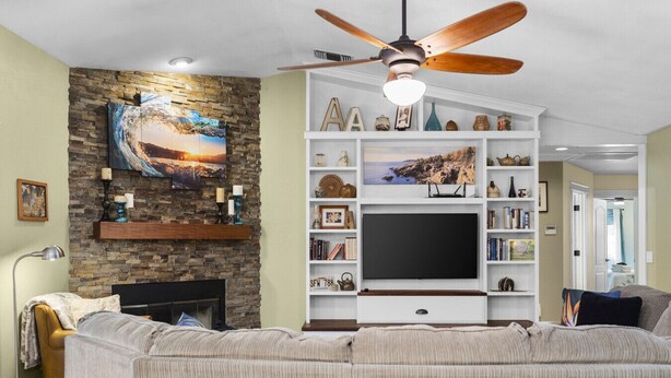 A living room with a fireplace, a large flat-screen TV, a ceiling fan, and a stone wall. - Hawaiian Coffee Hale (Redding)