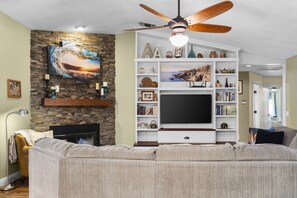 A living room with a fireplace, a large flat-screen TV, a ceiling fan, and a stone wall. - Hawaiian Coffee Hale | Central Home Minutes From Bethel (Redding)