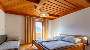Habitació amb sostre de fusta, un llit amb capçalera, una tauleta de nit, una finestra amb cortines i vistes a les muntanyes.