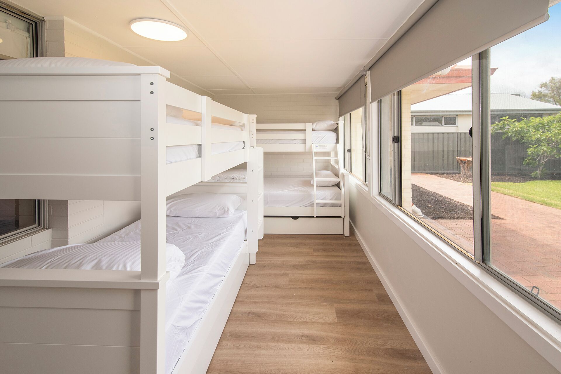 A narrow room with bunk beds, wooden flooring, and a window with a view of a backyard.