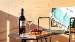 A bottle of Zweigelt wine, two glasses, and a wooden board with grapes and crackers on a round table in a room with a wooden wall and a chair with a green blanket.