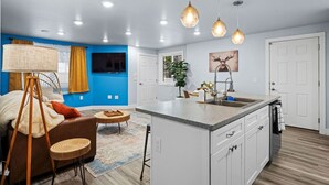 Une cuisine moderne dotée d’un îlot central, un coin repas avec un canapé et des chaises, et un salon avec un téléviseur et un mur d’accent bleu.