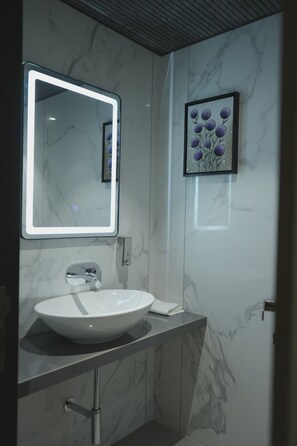 A bathroom with a white sink, a mirror with a light, and a framed artwork on the wall.