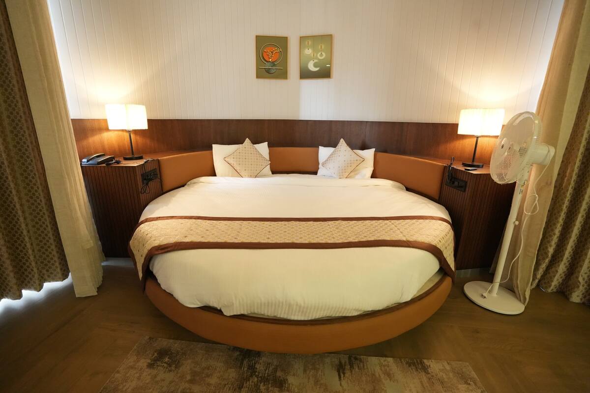 Una habitación de hotel con una cama grande, dos mesitas de noche con lámparas, una mesita con teléfono y un ventilador de pared.