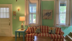 Une pièce avec un mur vert, un canapé à carreaux, une petite table avec une lampe, une carte postale accrochée au mur et une fenêtre avec des rideaux.
