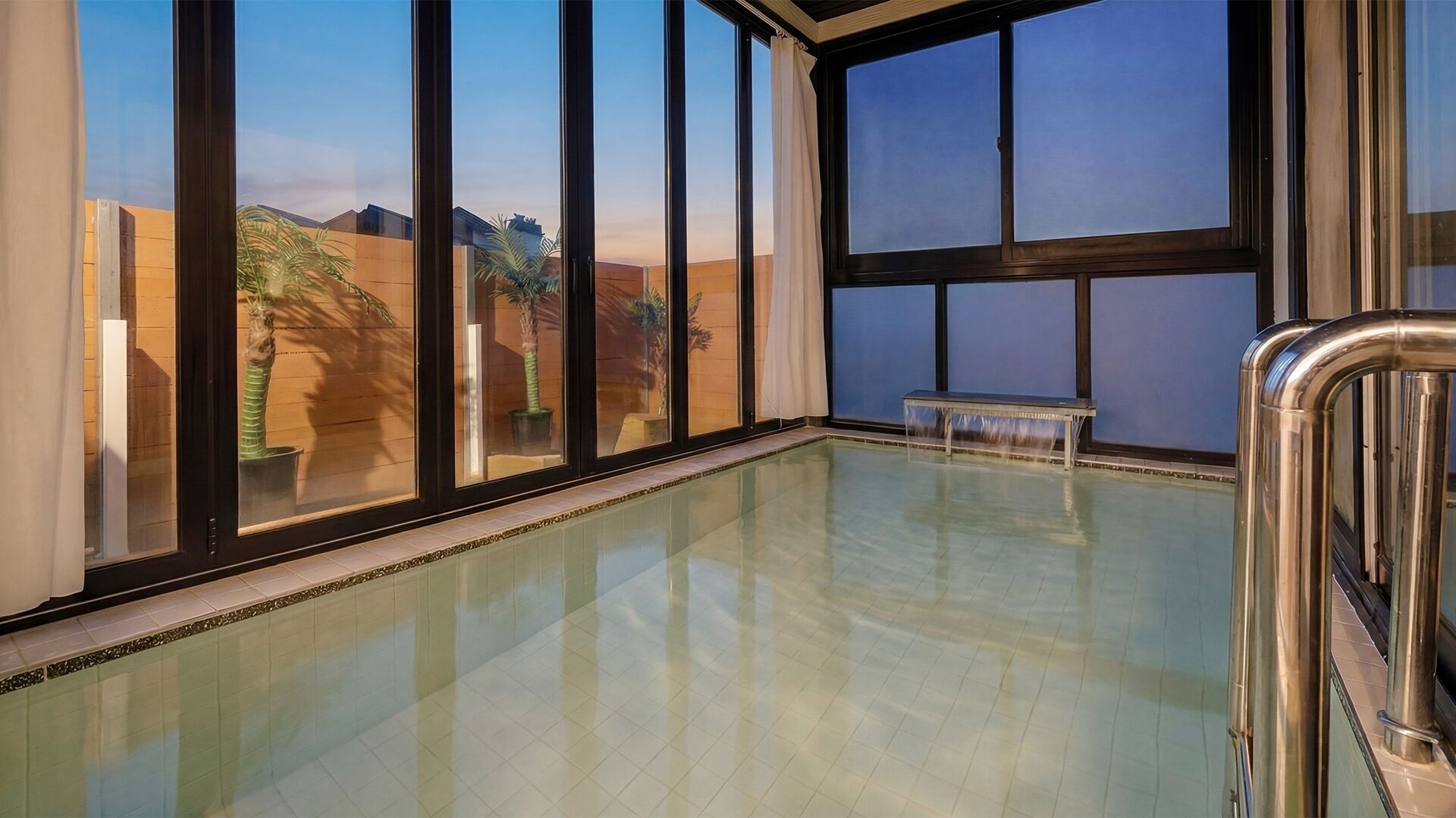 A spacious indoor swimming pool with large windows, a bench, and a metal railing.