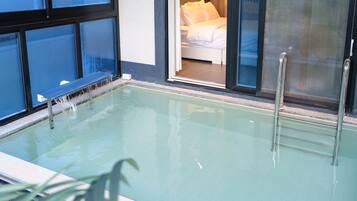 An indoor swimming pool with clear water, a glass barrier, and a view of a bedroom with a bed and pillows.