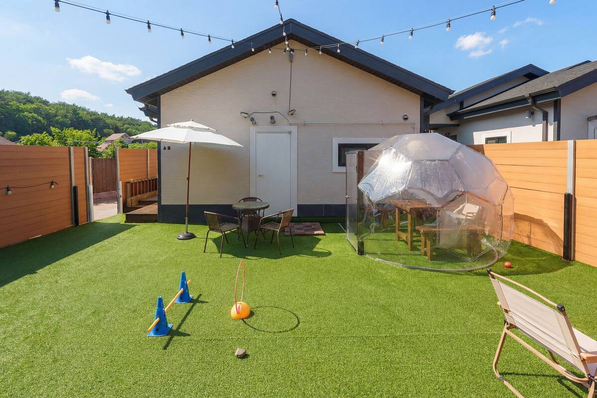 A backyard with artificial grass, a transparent dome structure, a wooden fence, and a house with a sloped roof.