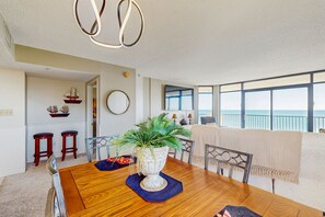 A dining area with a wooden table, a potted plant, and a view of the ocean through large windows. - Oceanfront condo w/private balcony, pool, direct beach access & close to golf (North Myrtle Beach)