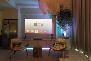 Ein modernes Wohnzimmer mit einem großen Apple TV Bildschirm, zwei Sesseln, einem kleinen runden Hocker und einer Topfpflanze.