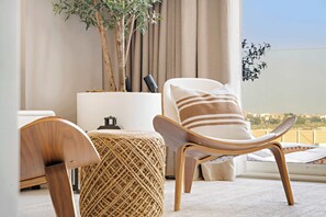 A modern living room with a wooden chair, a round ottoman, and a white sofa with striped cushions. A potted plant and a lamp are on a side table. - Silkhaus Luxury 1BR | Mamsha Saadiyat Sea View (Abu Dhabi)
