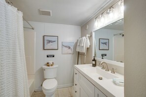 Baño con inodoro, lavabo y espejo. Hay cuadros enmarcados en la pared y un perchero para toallas.