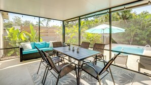 Een zonnig terras met een tafel en stoelen, een bank en uitzicht op een zwembad en groen.