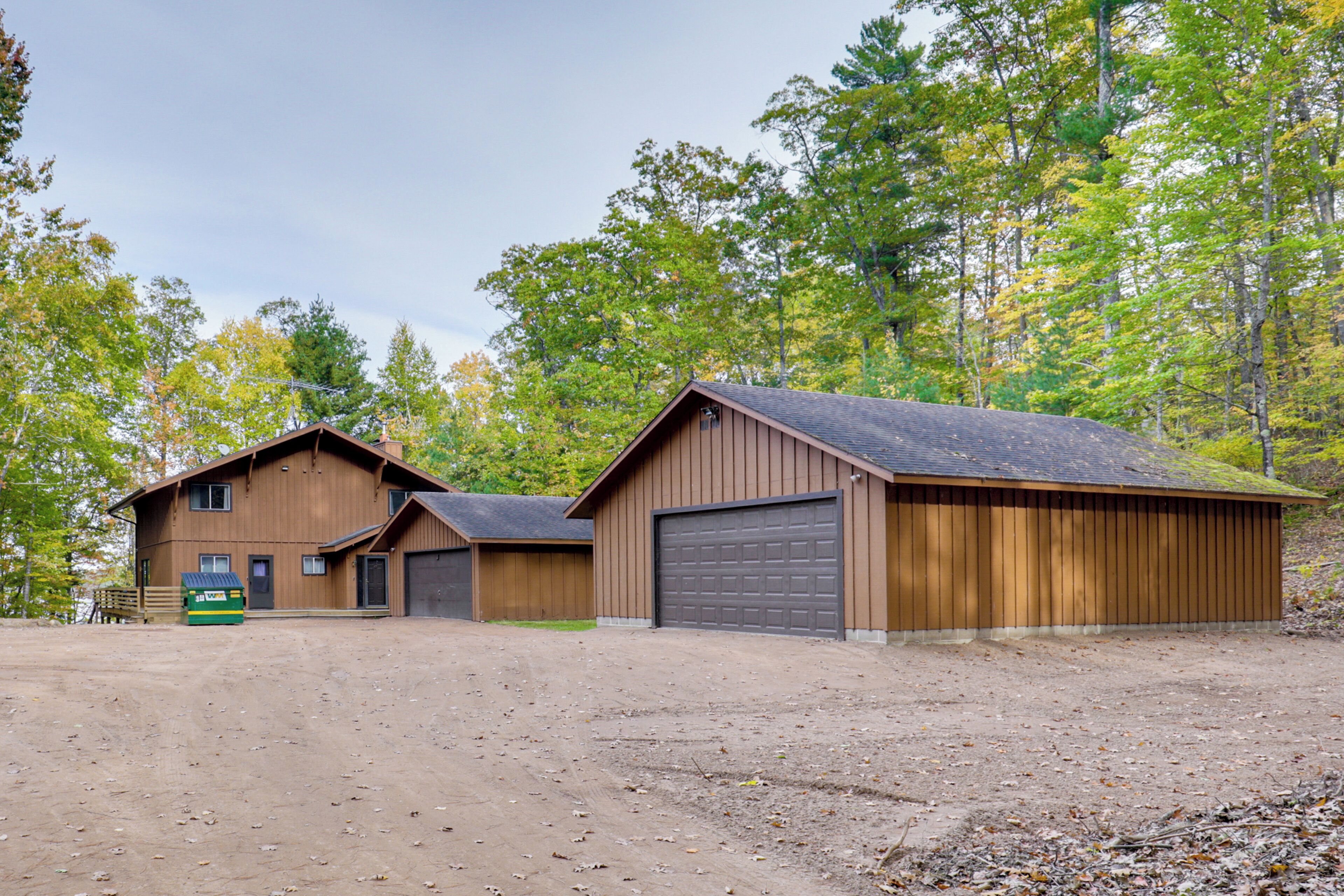 Une construction en bois à deux étages, dotée d’un garage, entourée d’arbres et bordée d’une allée non pavée.
