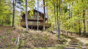 A wooden cabin with a balcony, surrounded by trees and a stone staircase leading up to it. - Land O' Lakes Cabin on Big Portage w/ Fire Pit (Land O' Lakes)