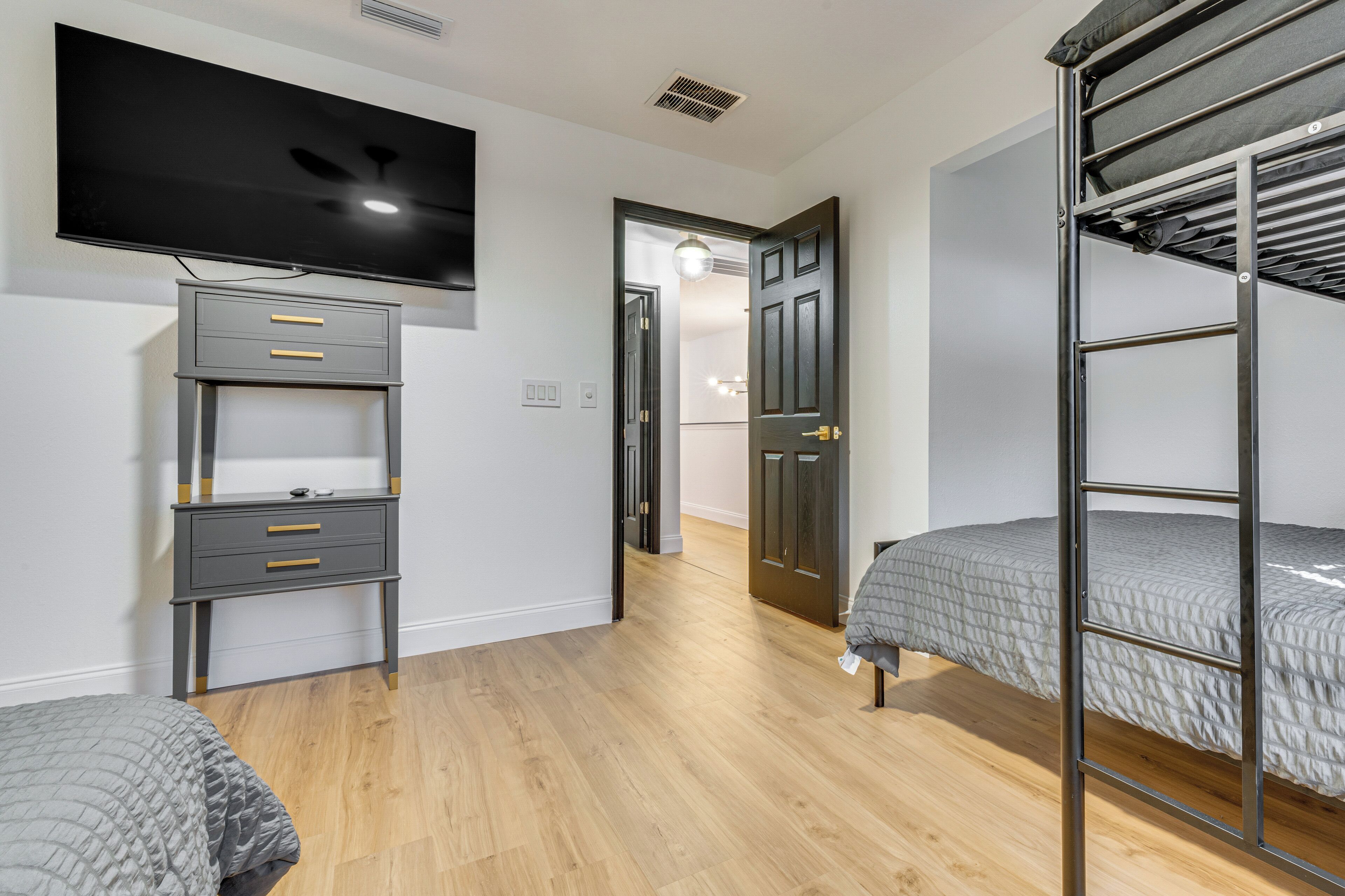 Un dormitorio con literas, un televisor montado en la pared, un escritorio con cajones y una puerta que da a otra habitación.