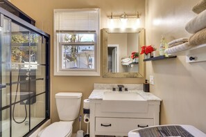 Et badeværelse med et glasvæg-afskærmet brusebad, toilet, håndvask med et spejl over, og et håndklædestativ med sammenfoldede håndklæder.