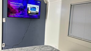 Un dormitorio con una televisión instalada en la pared mostrando una pantalla de actualización del sistema operativo, una colcha estampada y un control remoto sobre la cama.