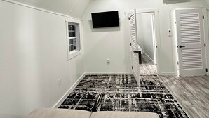 Una habitación con ventilador de techo, televisor montado, ventana con persianas, puerta con diseño de listones y alfombra estampada.
