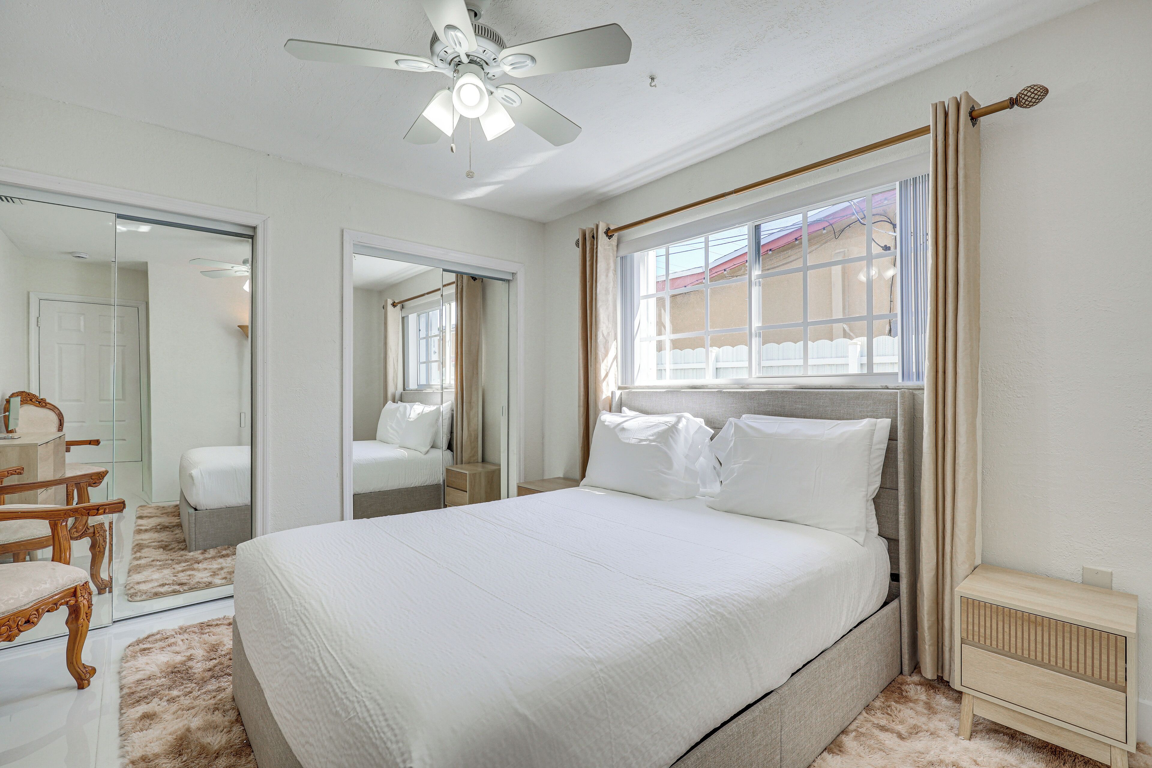 Quarto com cama grande, ventilador de teto, janela com cortinas, mesa de cabeceira e espelho.