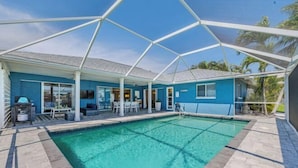 A pool area with a clear blue pool, a covered seating area, and a view of palm trees. - Cape Coastal (Cape Coral)