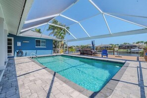 A swimming pool with a covered area, a blue building, and a view of palm trees and houses in the background. - Cape Coastal (Cape Coral)