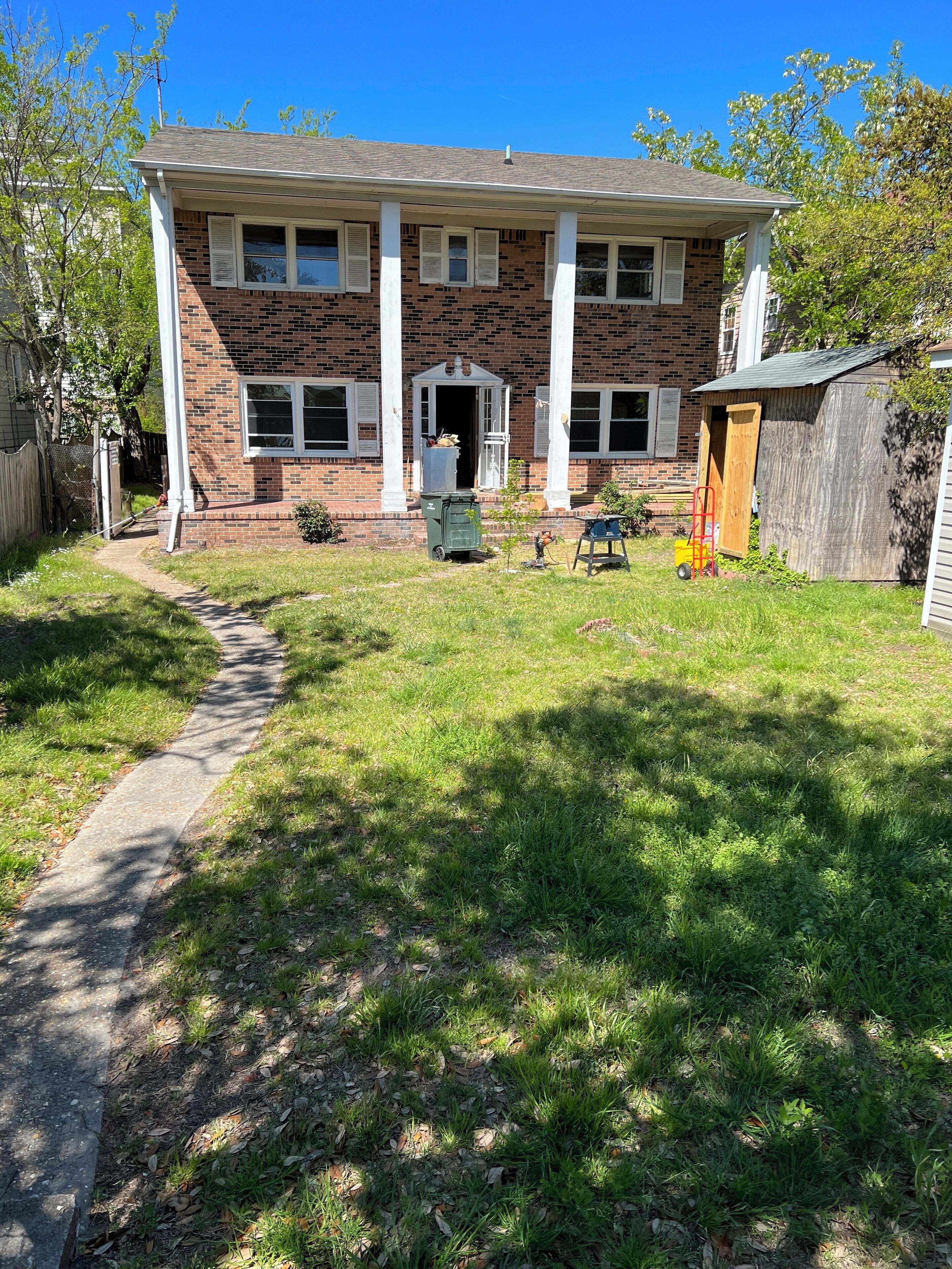 A two-story brick house with a front porch, a small shed, and a pathway leading to the front door.