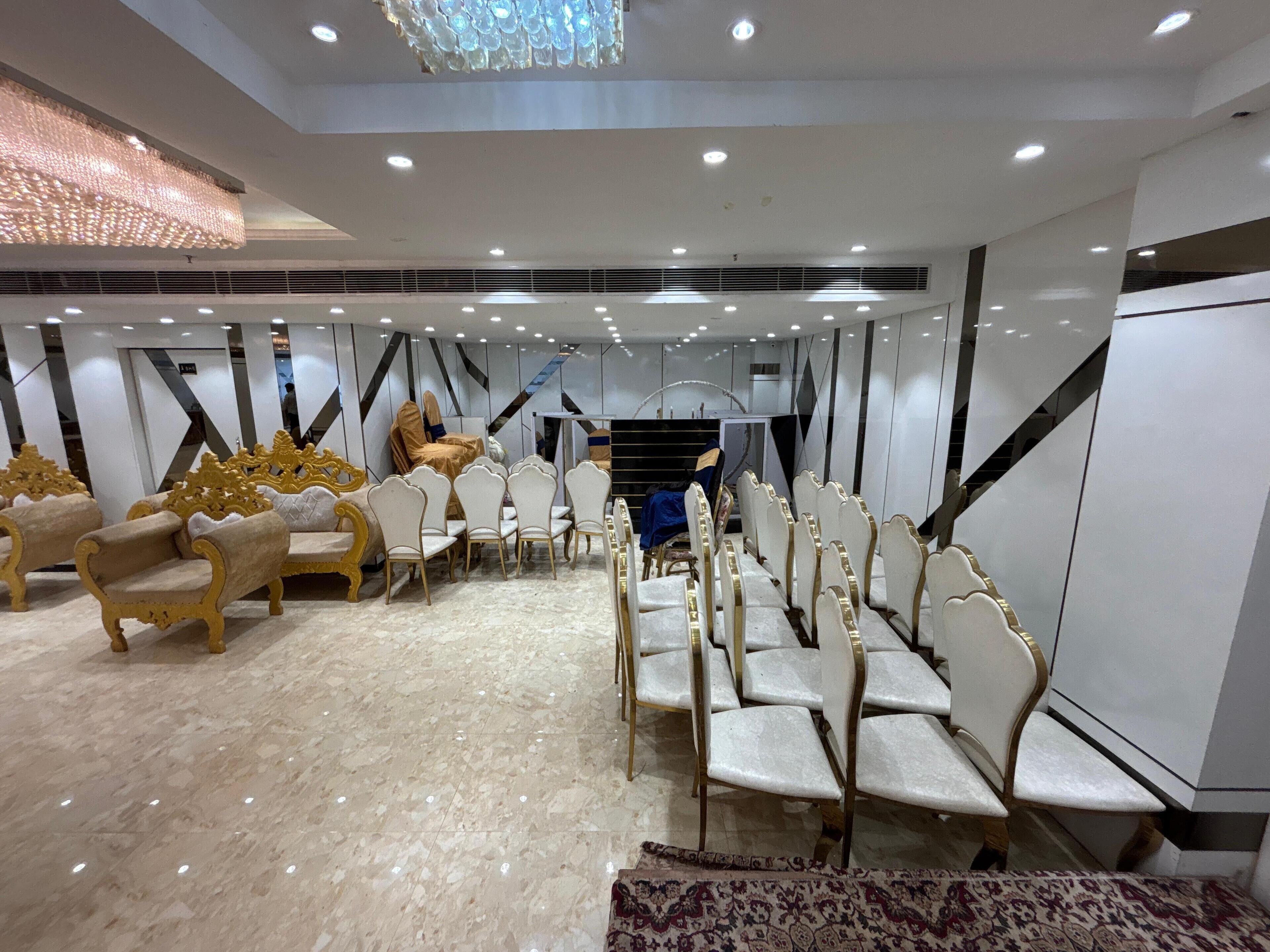 A spacious hall with rows of white chairs and golden chairs, a patterned rug, and a staircase in the background.