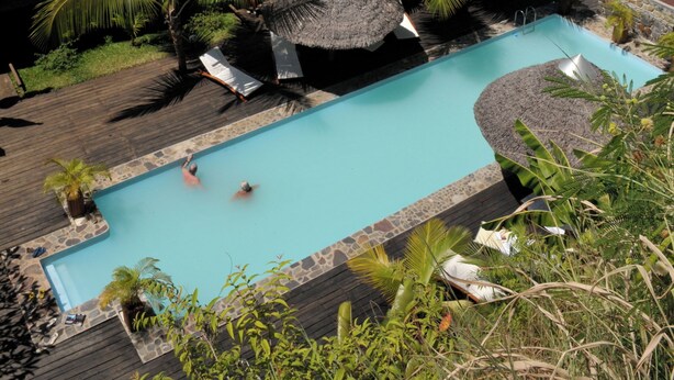 Zona de piscina con vista al océano, rodeada de palmeras y construcciones de techo de paja.