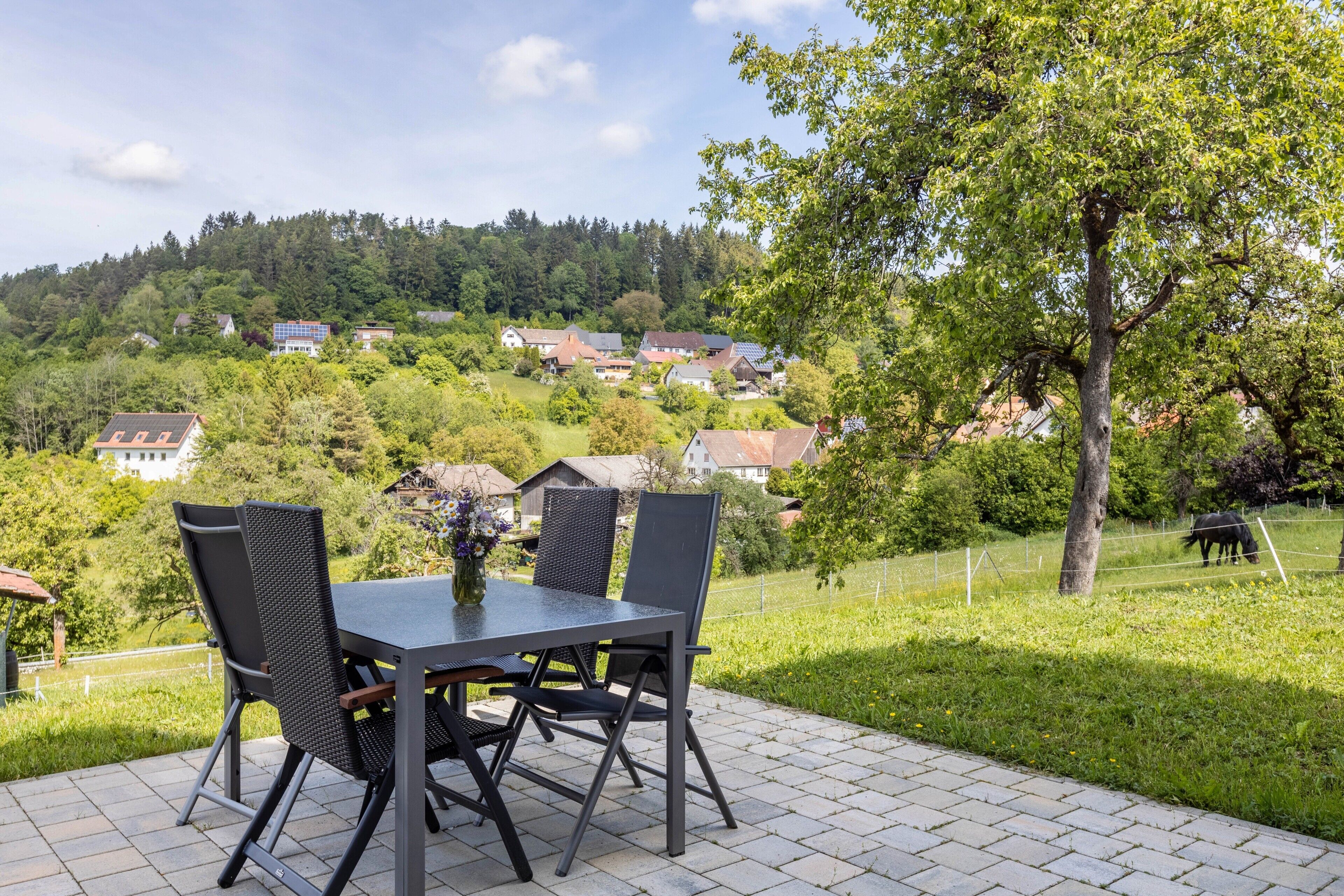 Une terrasse avec une table et des chaises, un arbre, et une vue sur des maisons au loin.