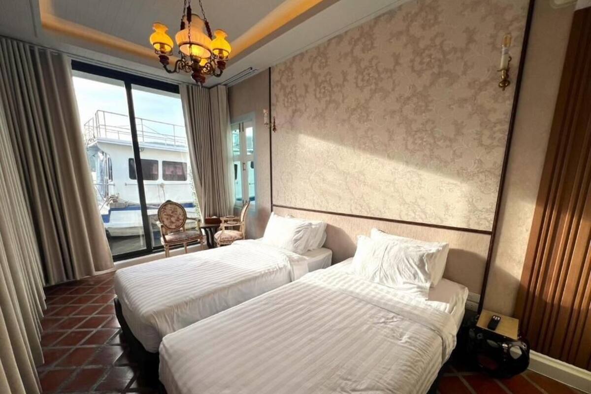 A hotel room with two beds, a chandelier, a large window with curtains, and a view of a boat outside.