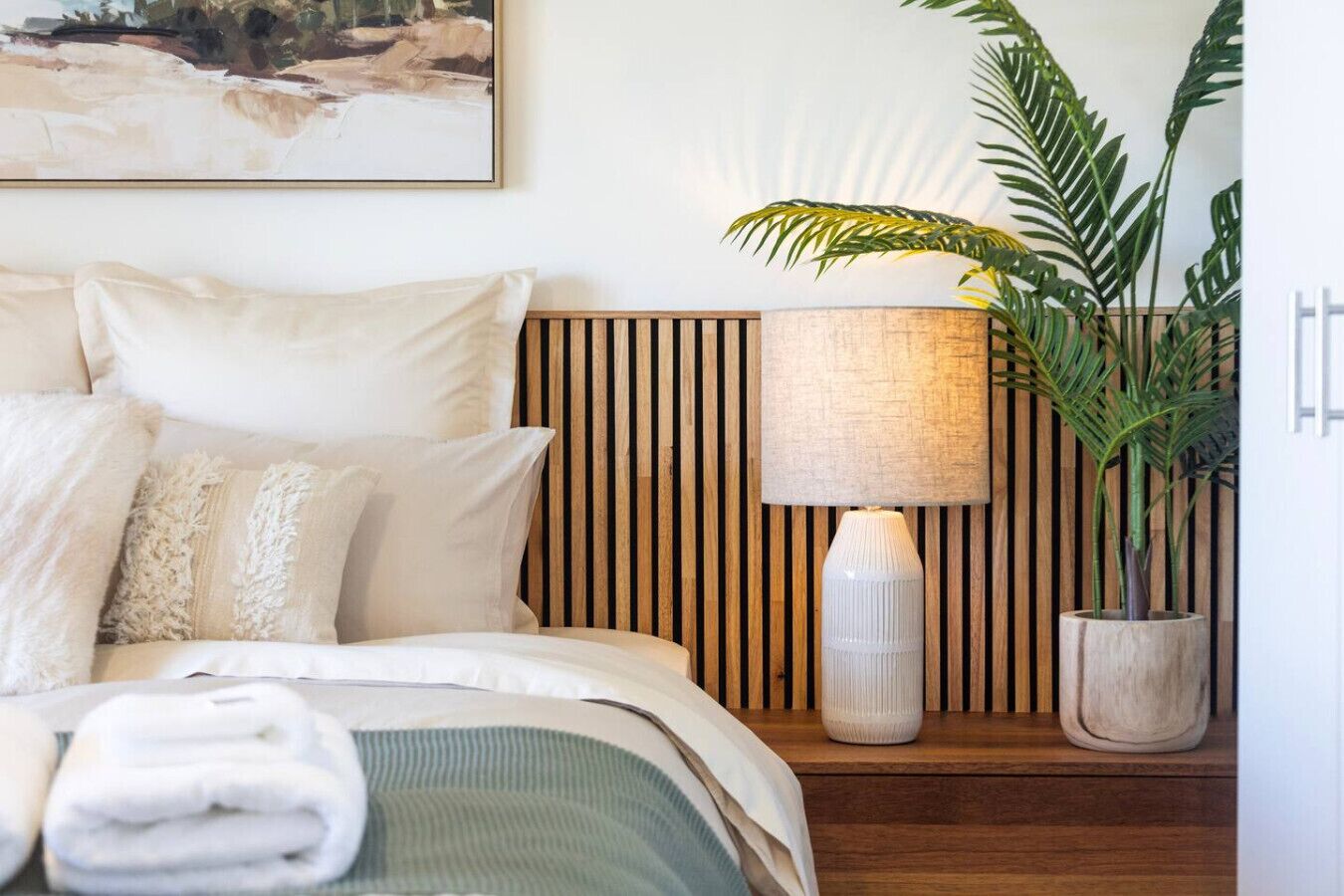 A neatly made bed with a green blanket, a wooden slatted headboard, a white lamp, and a potted plant.