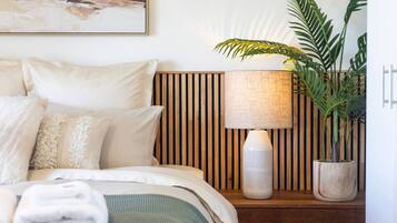 A neatly made bed with a green blanket, a wooden slatted headboard, a white lamp, and a potted plant.