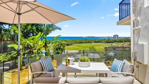 Une terrasse extérieure avec une table et des chaises, un parasol beige et une vue sur la mer et la verdure.