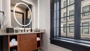 Ein modernes Badezimmer mit Waschtisch, einem beleuchteten Spiegel, einem Fenster und Blick auf Gebäude.