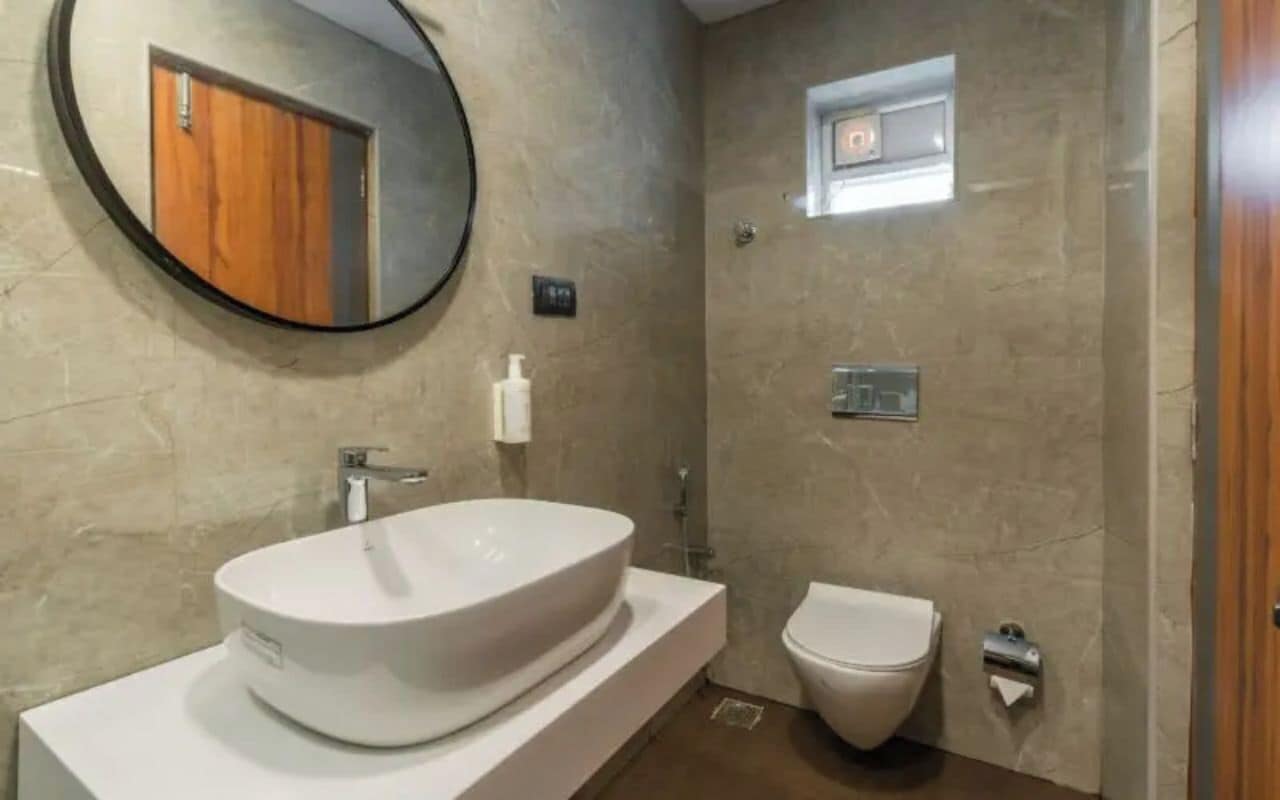 Une salle de bain avec un grand lavabo ovale, des toilettes, une petite fenêtre et un miroir qui reflète la porte en bois.