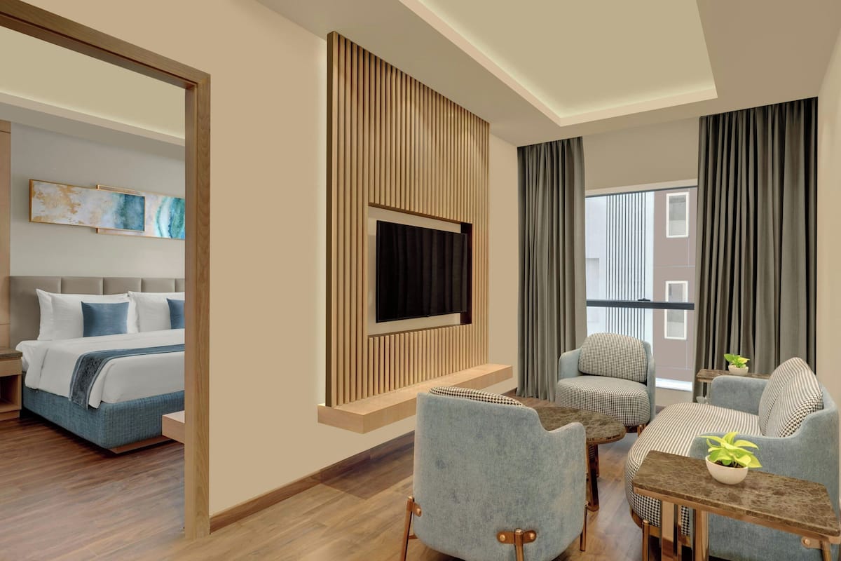 A modern hotel room with a bed, a TV mounted on a wooden panel, a seating area with a table and chairs, and a large window with curtains.