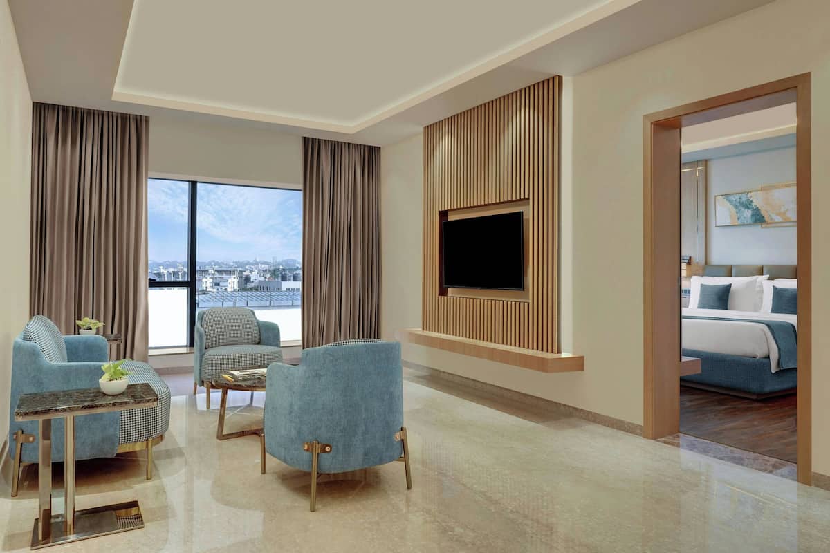 A modern hotel room with a large window, a flat-screen TV mounted on a wooden panel, a bed with blue bedding, and a seating area with blue armchairs and a small table.