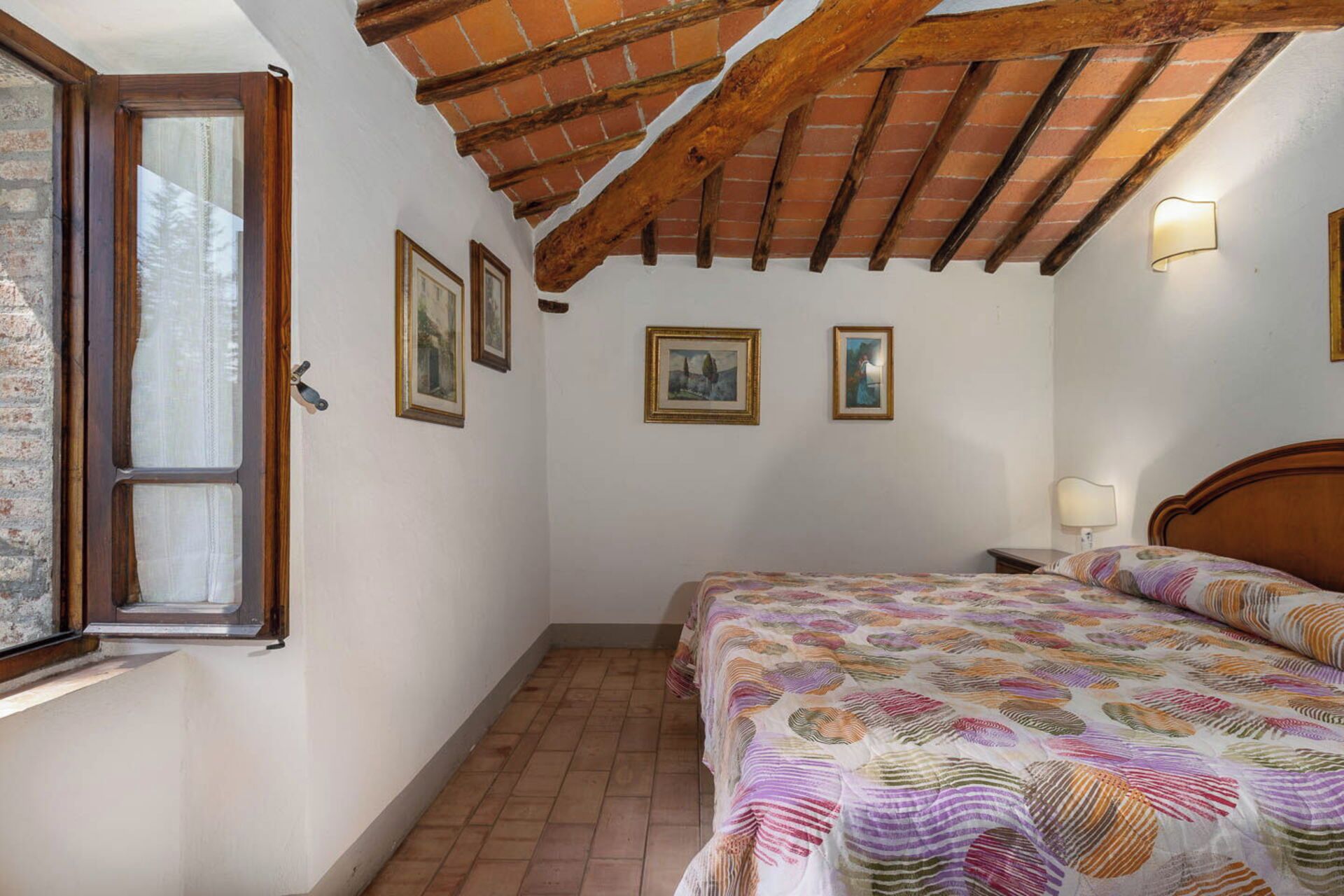 Une chambre avec un plafond en bois, un lit recouvert d’une couvre-lit à motifs, une table de chevet avec une lampe et des tableaux encadrés accrochés au mur.