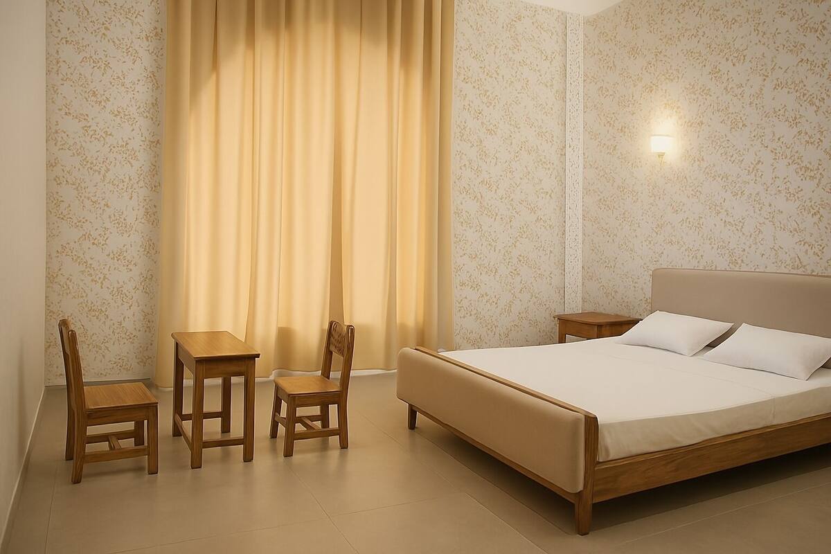 A bedroom with a bed, two chairs, a small table, a nightstand, and a window with curtains.