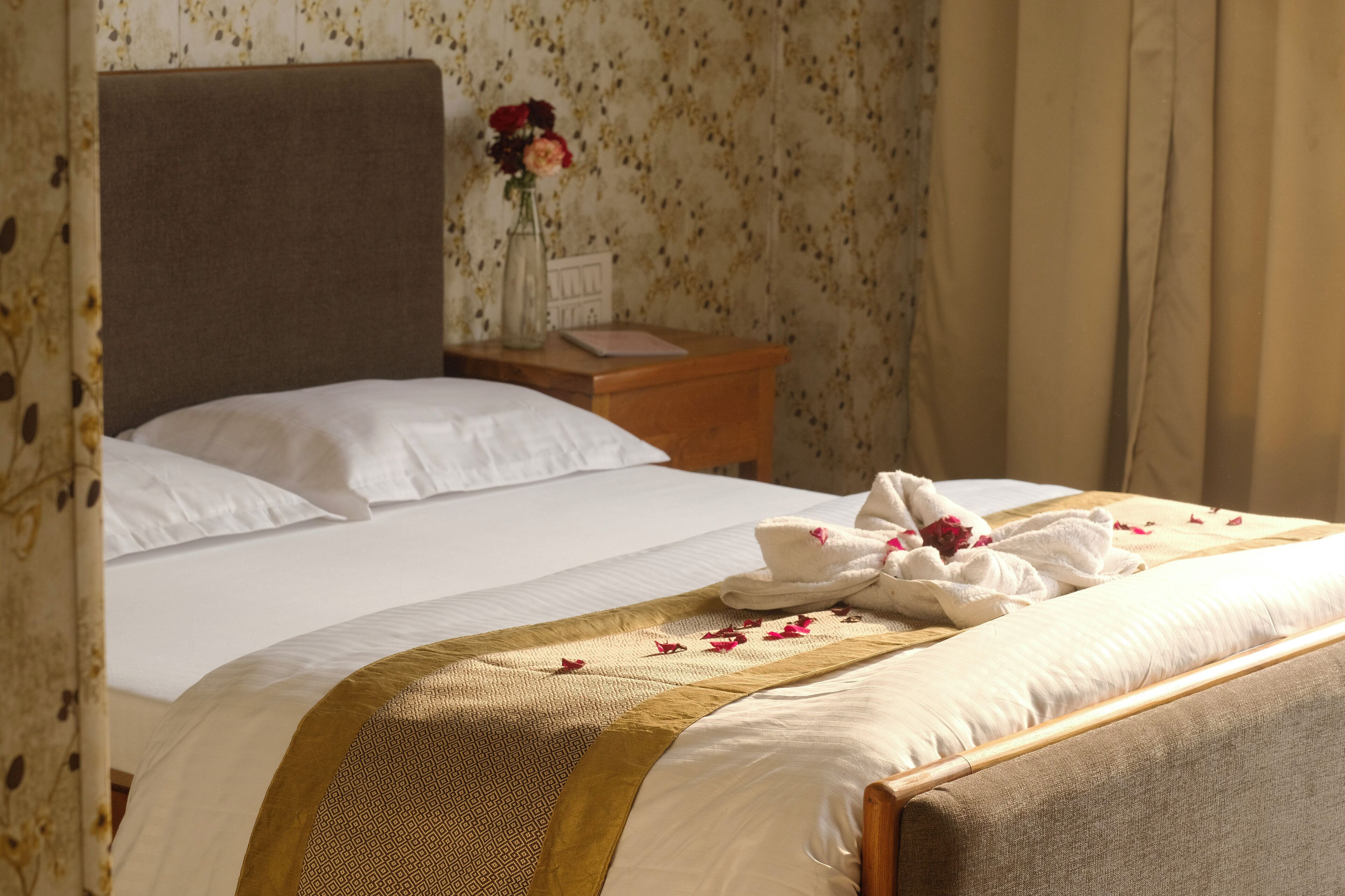 A neatly made bed with white linens and a decorative gold and brown bedspread. There is a wooden nightstand with a vase of flowers and a book beside the bed.