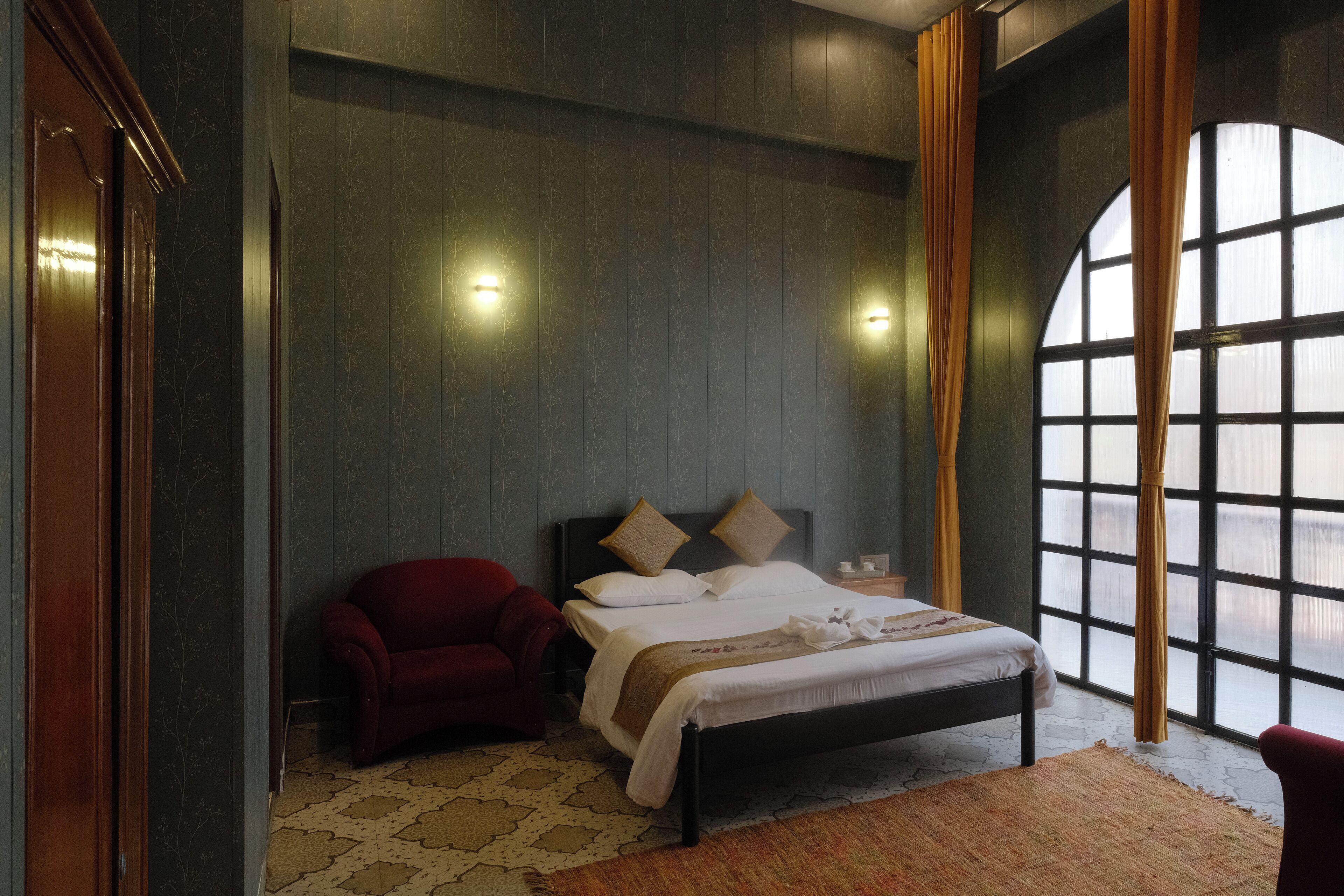 A hotel room with a large arched window, a bed with a headboard, a red armchair, and a patterned rug.