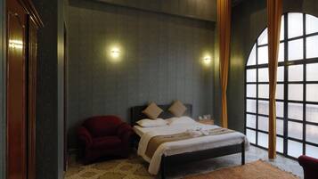 A hotel room with a large arched window, a bed with a headboard, a red armchair, and a patterned rug.
