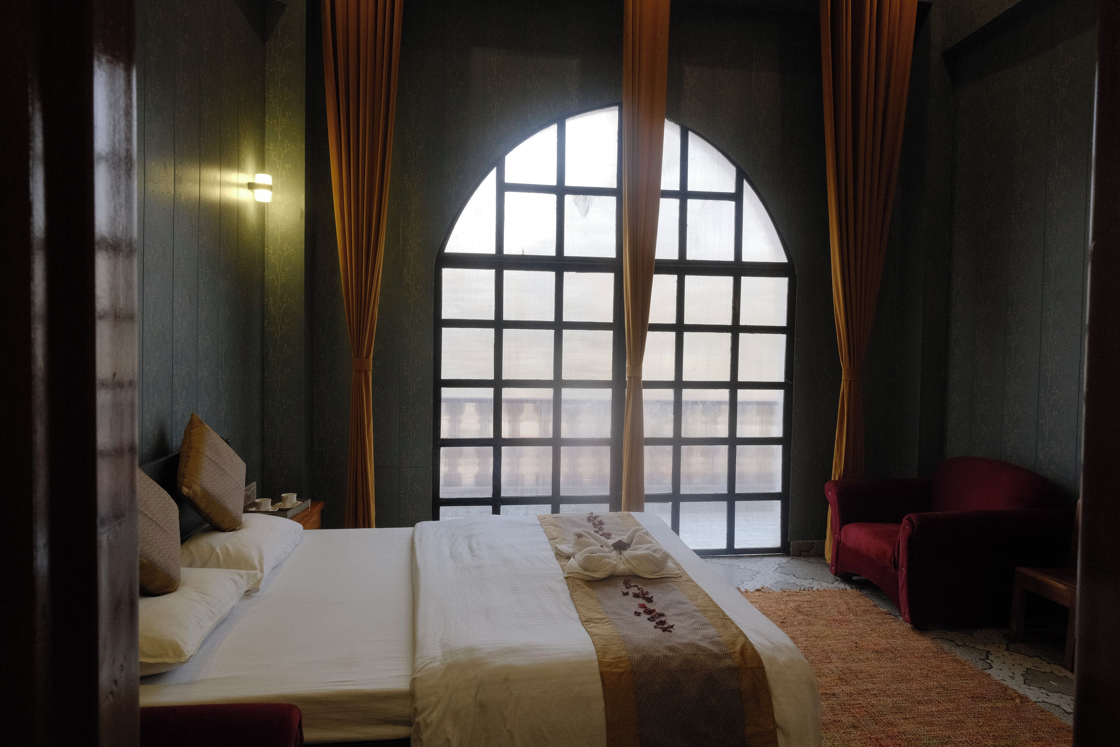 A hotel room with a large arched window, a bed with white linens, a red armchair, and a small table with a lamp.